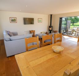 A living room with wooden dining table and chairs a gray sofa a wall-mounted TV a stove and an open door leading to an outdoor seating area at Little Owl Barn in Upton Snodsbury