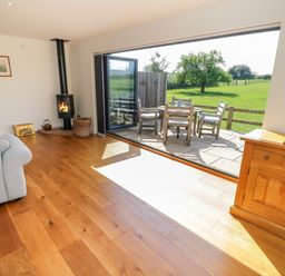 A living room with wooden floor a wood stove a gray sofa and a wooden cabinet opening onto a patio with outdoor furniture and green fields at Little Owl Barn in Upton Snodsbury