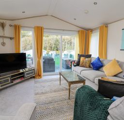 A living room with a grey sofa with blue and yellow cushions a patterned rug a glass coffee table a black TV on a black and gold TV stand and yellow curtains at 95 Hafan Taffy in Gilfachrheda near Llanarth