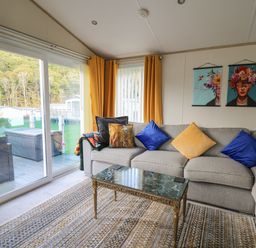 A living room with a gray sectional sofa with blue and yellow pillows a marble top coffee table orange curtains and sliding glass doors at 95 Hafan Taffy in Gilfachrheda near Llanarth