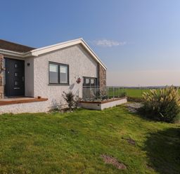 A house exterior with stone and white walls a black door and a glass balcony overlooking a grassy field at Corrie Knowe in Trearddur Bay