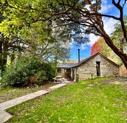 A stone house with a pitched roof surrounded by trees and a grass lawn with a paved pathway leading to the entrance at Harvey's House in Helston