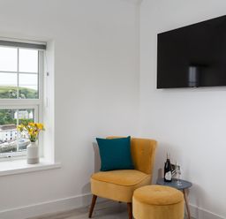 A corner with a yellow chair and ottoman a side table with a wine bottle and glass a window with flowers in a vase and a wall-mounted TV at Chy Lowen in Porthleven