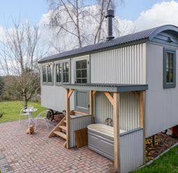 An outdoor patio with a small white table and chairs a wooden hut with corrugated metal sides and a covered bathtub on a paved area at Evendine Hut @ Evendine Cottage in Colwall Herefordshire