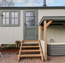 An exterior view of a corrugated metal building with green framed windows and door wooden steps leading to the door a covered outdoor grey bathtub and a white metal chair on a paved patio at Evendine Hut @ Evendine Cottage in Colwall Herefordshire