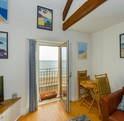 A room with a small dining table and chairs a leather sofa and an open door with a view of the sea at 2 Bayview in Torcross
