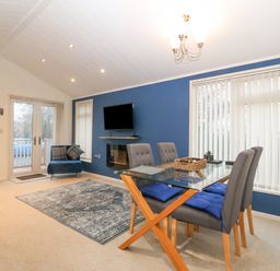 A dining area with a glass table and four chairs near a living space with a blue accent wall and a mounted tv at Y Caban in Brynsiencyn