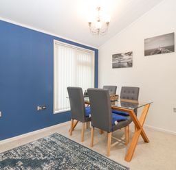 A dining room with a glass table and four grey chairs near a window with vertical blinds at Y Caban in Brynsiencyn