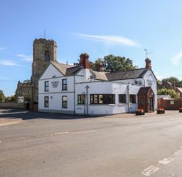 A white building with a sign reading Five Bells and an old church tower behind it at The Five Bells Inn in Upwell