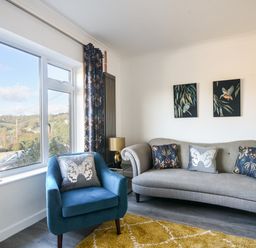 A living room with a gray sofa and a blue armchair near a large window with patterned curtains at Hook Farm Cottage in Uplyme