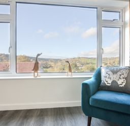 A window with bird figurines on the sill and a blue armchair with a butterfly pillow at Hook Farm Cottage in Uplyme