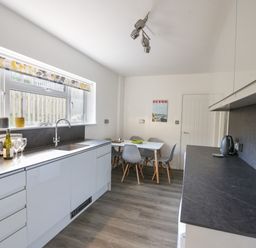 A kitchen with white cabinets gray countertops a sink window with patterned blind and a dining table with chairs at Hook Farm Cottage in Uplyme