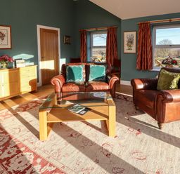 A living room with leather armchairs wooden coffee table sideboard and windows with red curtains at Summerseats Cottage in Alnwick