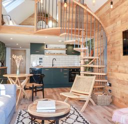 A living room with wooden walls spiral staircase and kitchen area at Little Beach Cabin in Crantock