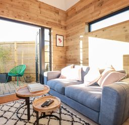 A living area with a gray sofa and pillows a patterned rug and nested wooden tables opening to a patio with green chairs at Little Beach Cabin in Crantock
