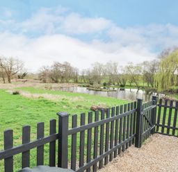 A black wooden fence with a gate overlooking a grassy area and a pond with trees at The Hayloft in Haddiscoe