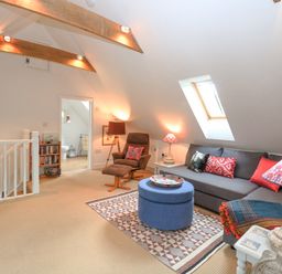 An attic lounge with a brown armchair a grey sofa with cushions and a blue ottoman on a patterned rug at The Hayloft in Haddiscoe