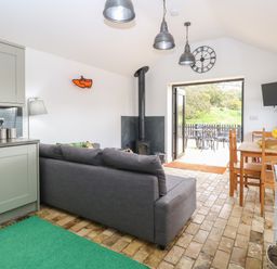 A living area with a grey sofa a wood stove a wooden dining table and chairs a wall clock and a TV near open doors leading outside at The Grain Store in Haddiscoe