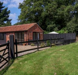 A small brick and wood building with a tiled roof near a fenced gravel area and green lawn at The Grain Store in Haddiscoe
