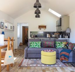 A living area with a gray sofa and colorful cushions next to a small kitchen and dining table at The Grain Store in Haddiscoe