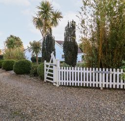 A white wooden picket fence gate along a gravel driveway with round bushes and tall trees at Lampland House in Walberswick