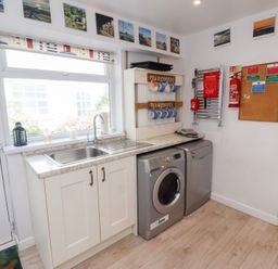 A utility room with a sink washing machine dishwasher and mugs on a rack at Seascape Cottage in Seahouses