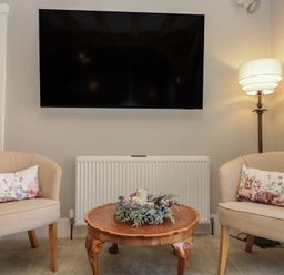 A sitting area with two beige chairs with animal print cushions a wooden coffee table with candles and flowers a floor lamp and a wall mounted television at The Barn Helperby near Boroughbridge