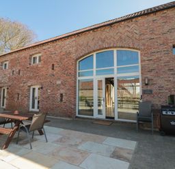An outdoor patio area with wooden picnic table and chairs next to a brick building with large arched window and doors at The Barn in Helperby near Boroughbridge