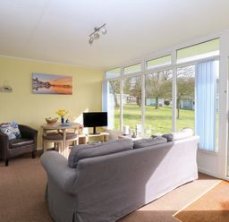 A living room with a gray sofa a round wooden table a leather chair and large windows overlooking a grassy area at The Beach Hut in Cromer