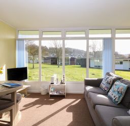 A living room with a gray sofa with decorative pillows a small wooden table with flowers a TV and large windows showing a lawn at The Beach Hut in Cromer