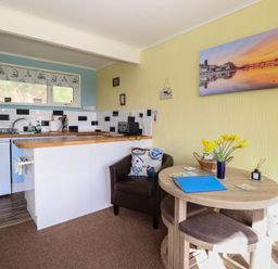 A kitchen area with blue cabinets a small fridge a round wooden table with chairs and a leather chair with cushions at The Beach Hut in Cromer