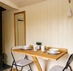 A small dining area with a wooden table set for two and a black wood stove at Mitford Gypsy Hut in Moreton-In-Marsh
