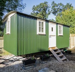 A green corrugated metal hut with white windows and door with wooden steps in a fenced garden area at Mitford Gypsy Hut in Moreton-In-Marsh
