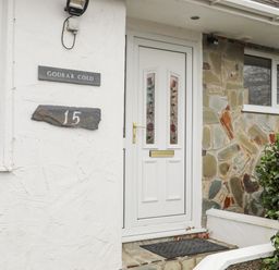 The entrance door of a house with a white door and stained glass panels next to a stone wall with house number 15 at 15 Maes Awel in Abersoch