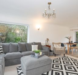 A living room with a grey sectional sofa and ottoman a beige armchair a dining table and chairs with sliding glass doors at 15 Maes Awel in Abersoch