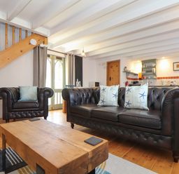 A living room with a leather sofa and armchair a wooden coffee table a staircase and a kitchen area at Oofoo's Barn in Mullion