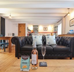 A living room with a black leather sofa and starfish cushions a wooden table with a bottle of wine a glass and a box of chocolates at Oofoo's Barn in Mullion