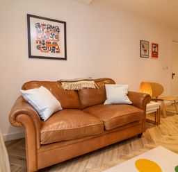 A brown leather sofa with two white pillows and a throw blanket in a room with framed artwork a wooden chair a side table with a lamp and wooden flooring at Valise in Bideford