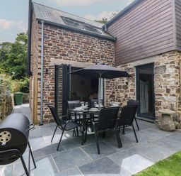 An outdoor patio with a dining table and chairs under an umbrella next to a stone house with glass doors and a barbecue grill at Wildwood in Newquay