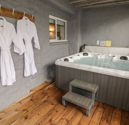 An indoor hot tub with water jets and a small step stool next to two white bathrobes hanging on hooks on a gray stone wall at Le Durkestrete Cellar in Haverfordwest