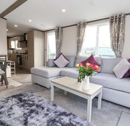 A living room with a gray sectional sofa and a coffee table with flowers near a kitchen and dining area at Ginger Fox Lodge in East Heslerton near Cayton