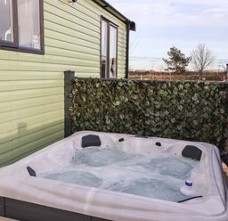 An outdoor hot tub with bubbling water next to a light green cabin with a privacy screen covered in artificial leaves at Ginger Fox Lodge in East Heslerton near Cayton