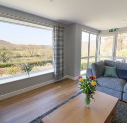 A living room with a grey sofa a wooden coffee table with flowers and large windows showing a countryside view at Curtau in Llanfaredd near Builth Wells