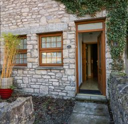 The exterior of a stone house with two windows and an open wooden door with ivy on the wall at Time and Tide in Arnside