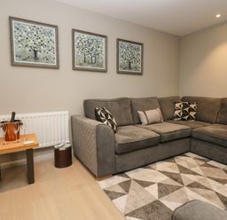 A living room with a grey sectional sofa a patterned rug three framed pictures on the wall and a small wooden table with wine glasses and a wine cooler at Brimstone in Backbarrow near Ulverston