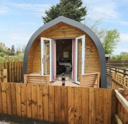 A wooden pod with open doors and a small patio table with chairs and wine glasses at The Poddy in Bushton near Lyneham