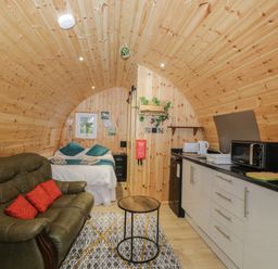 A wooden cabin interior with a bed green sofa small round table and kitchenette at The Poddy in Bushton near Lyneham