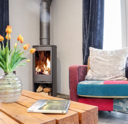 A living room with a vase of orange tulips on a wooden table a colorful armchair and a burning wood stove at La Petit Maison in St Columb Major