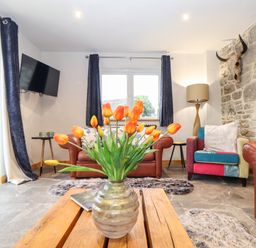 A living room with orange flowers on a wooden coffee table and leather sofas at La Petit Maison in St Columb Major