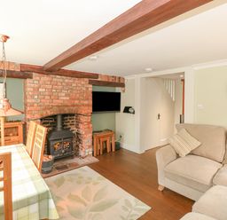 A living room with a brick fireplace, wood-burning stove, dining table with chairs, and a beige sofa at Easter Cottage in Dersingham